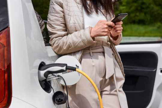 Young woman standing near the electric car with mobile phone in her hand and waiting for recharging of the automobile battery