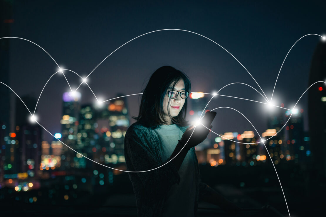 A woman looks at her smartphone and smiles, with a city skyline in the background and a connected network of lights superimposed on top.