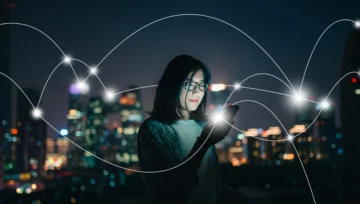 A woman looks at her smartphone and smiles, with a city skyline in the background and a connected network of lights superimposed on top.