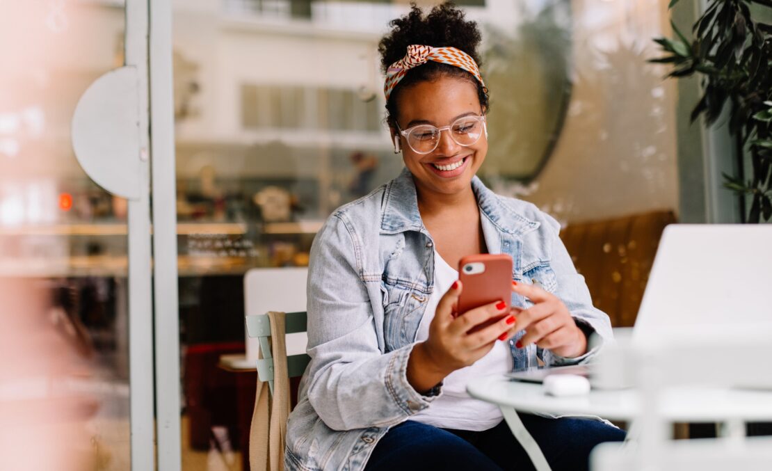 A customer sits in a cafe and smiles at her phone as she communicates with customer service.