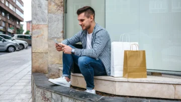 young man sitting outside on steps texting on his phone with shopping bags next to him
