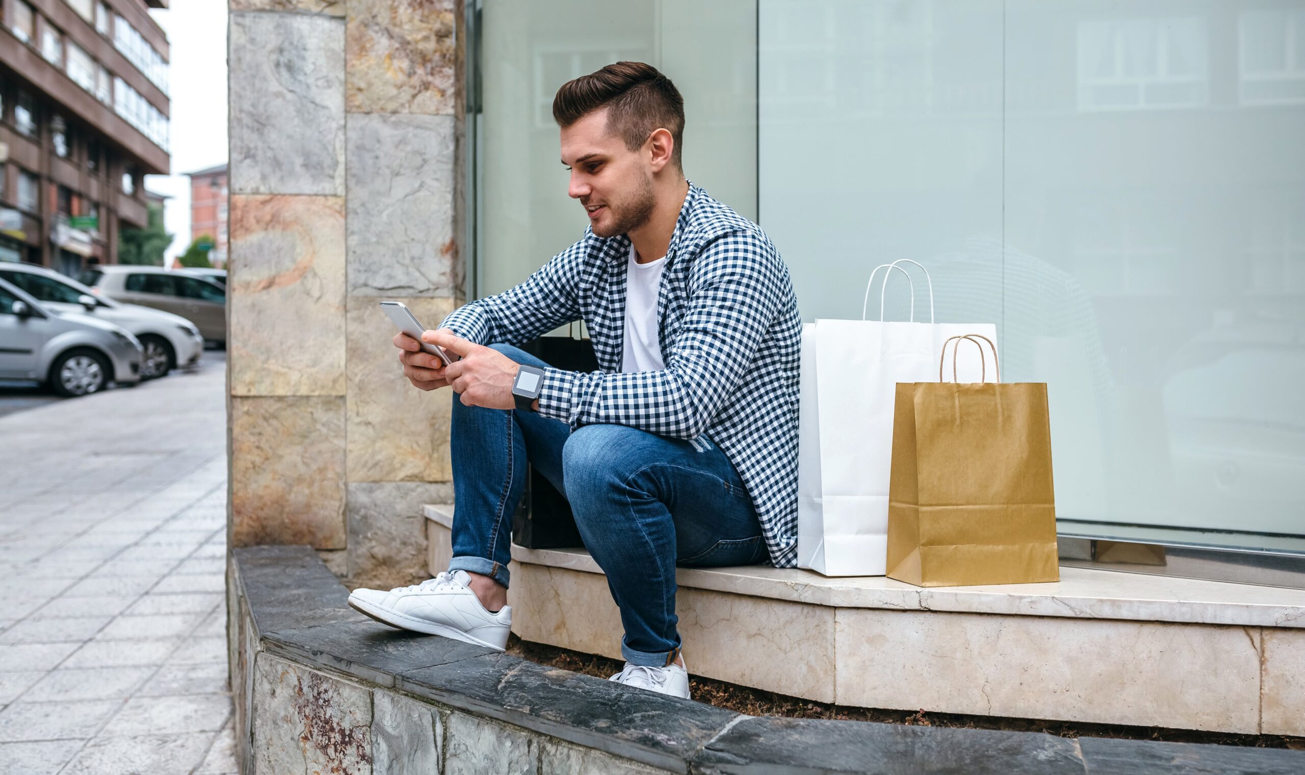 young man sitting outside on steps texting on his phone with shopping bags next to him
