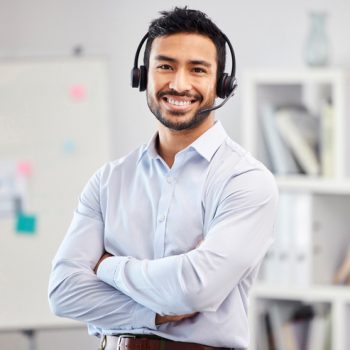 Young happy mixed race call center agent standing with his arms crossed in an office at work. One hispanic customer service agent wearing a headset and answering calls while standing in an office